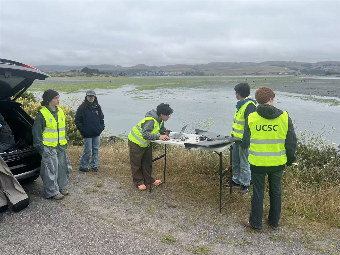 GeoFly Lab team in UCSC safety vests preparing a fixed-wing drone at a folding table beside an eelgrass mudflat at low tide